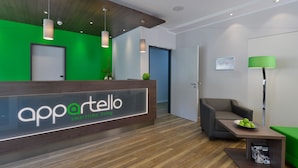 A reception area with a wooden reception desk, a green wall, and a modern lamp. - appartello - smarttime living Hamburg (Hamburg)