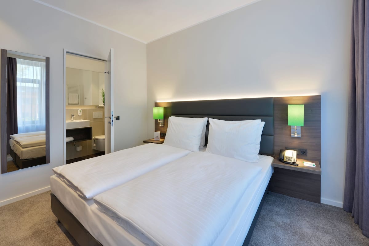 Une chambre d’hôtel avec un grand lit, une table de chevet avec une lampe, un miroir et une salle de bain visible à travers une porte ouverte.