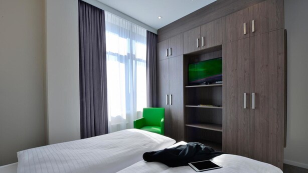 A hotel room with two beds, a green chair, and a TV on a wooden cabinet. - appartello - smarttime living Hamburg (Hamburg)