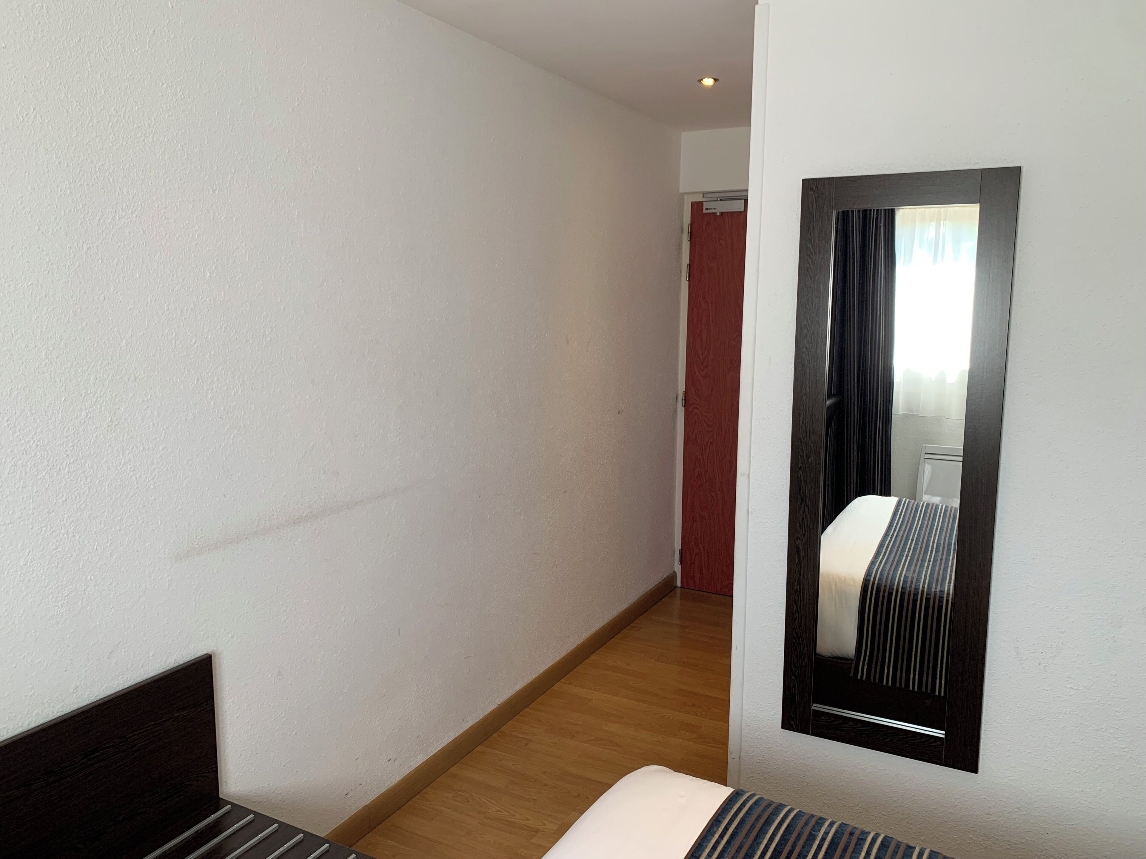 Habitación de hotel con cama, piso de madera, pared blanca y espejo que refleja una ventana con cortinas.