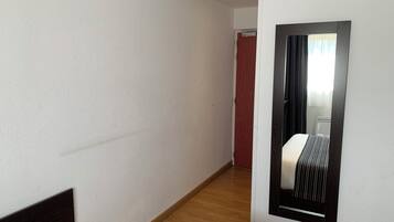 Habitación de hotel con cama, piso de madera, pared blanca y espejo que refleja una ventana con cortinas.