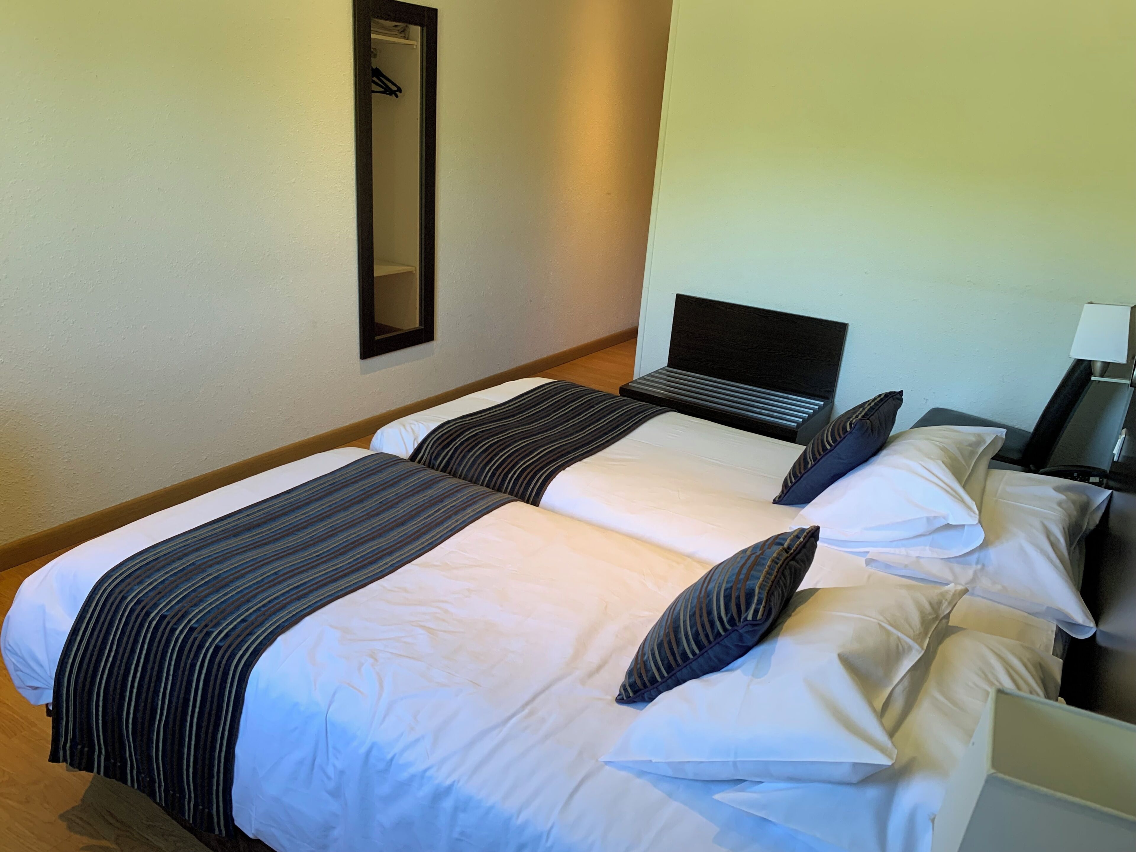 A hotel room with two beds, a nightstand, a lamp, and a mirror on the wall.