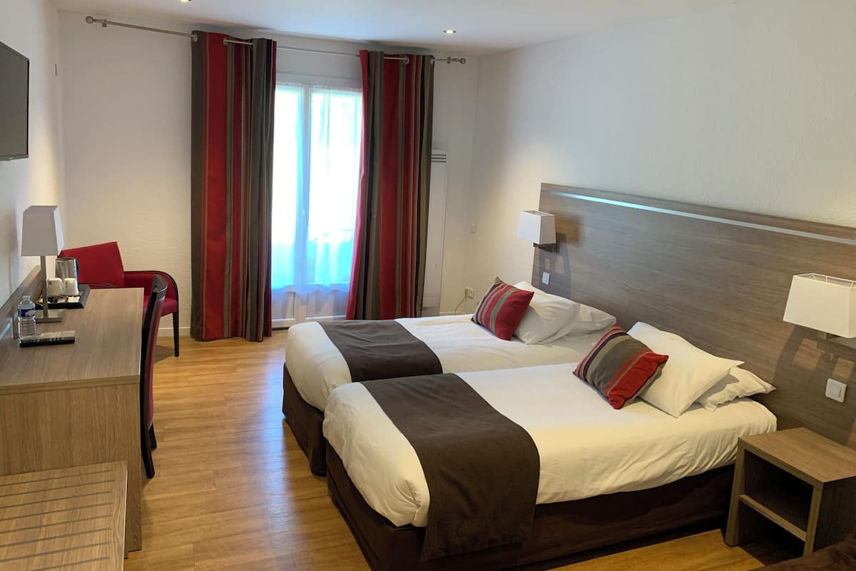 A hotel room with two beds, a desk with a chair, a bedside table, and a window with curtains.