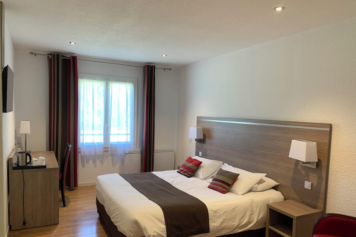 A hotel room with a bed, bedside table, lamp, and a desk with a lamp.