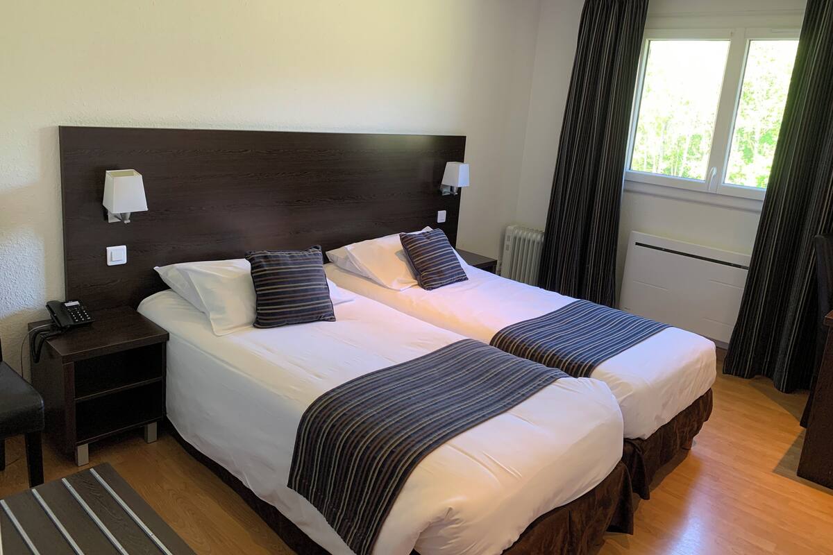 A hotel room with two beds, a nightstand with a phone, a lamp, a window with curtains, and a radiator.