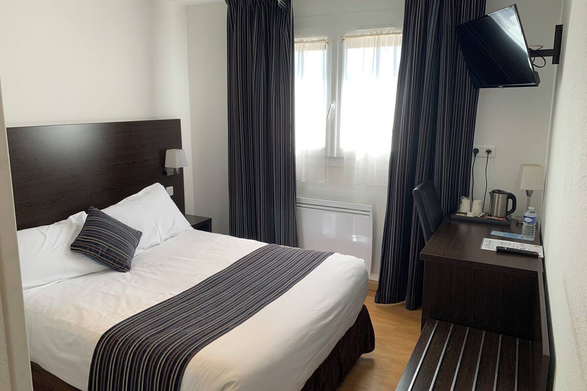 A hotel room with a bed, a TV, a bedside table with a lamp, a window with curtains, and a radiator.