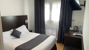 A hotel room with a bed, a TV, a bedside table with a lamp, a window with curtains, and a radiator.