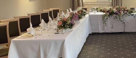 A banquet hall with long tables set for a formal event, featuring white tablecloths and floral centerpieces.