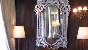 Una habitación con un espejo grande y ornamentado, dos lámparas, un jarrón con flores y un sillón blanco.