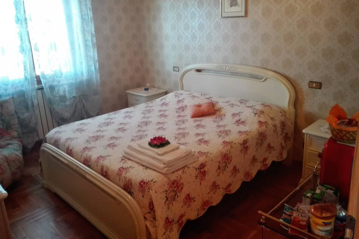 Een slaapkamer met een bed, nachtkastje, raam met gordijnen en een aan de muur hangend schilderij.