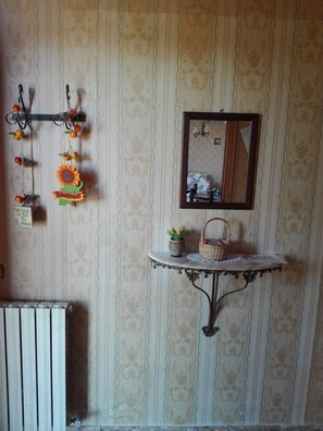 Une pièce avec un radiateur, un miroir fixé au mur, une petite table avec un panier et une plante en pot, et une décoration murale représentant un tournesol et un panneau indiquant « Bonjour ».
