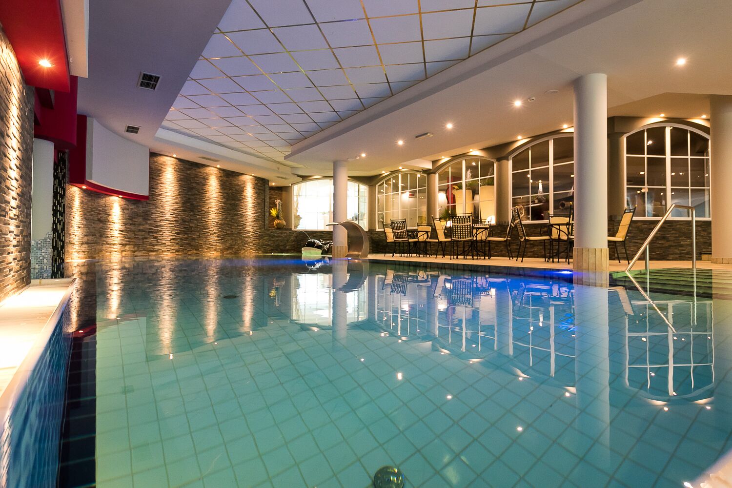 An indoor swimming pool with tiled walls, a glass ceiling, and seating areas with tables and chairs.