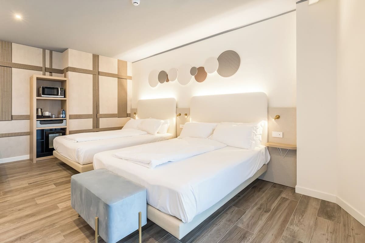 A modern hotel room with two beds, a built-in microwave, a small kitchenette, and a wall-mounted light fixture.