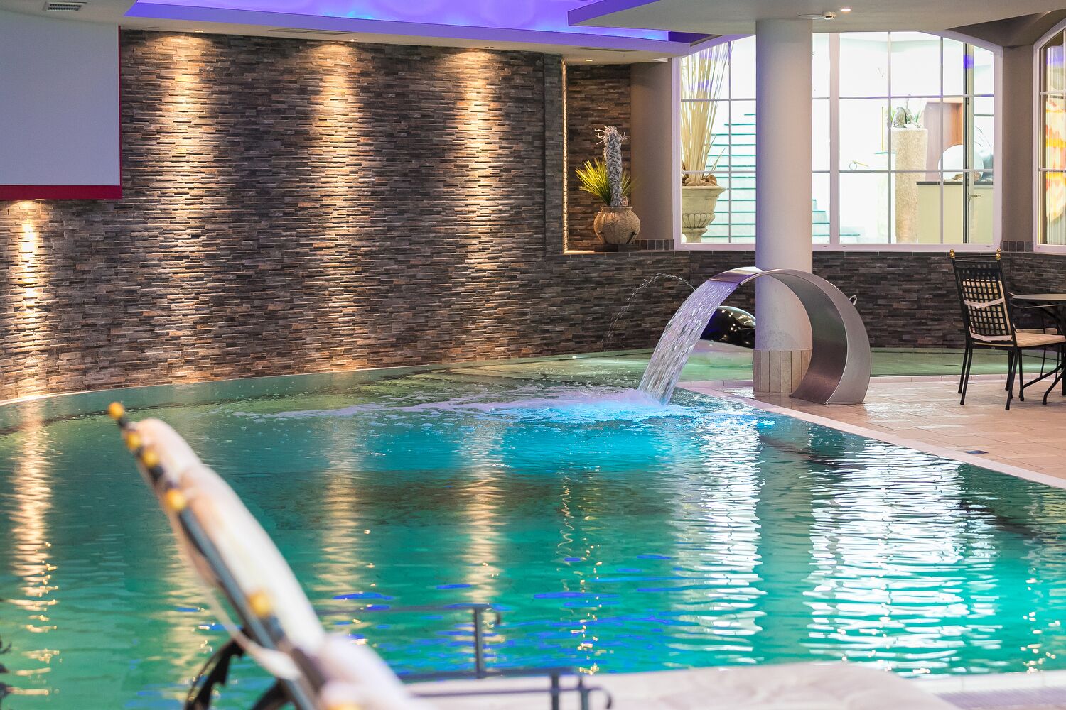 A swimming pool with a waterfall feature, surrounded by stone walls and indoor seating.