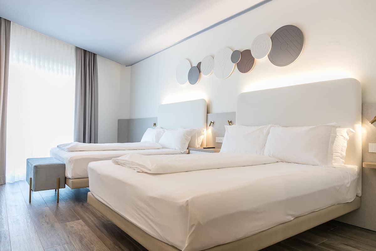 A hotel room with two beds, a bedside table, a chair, and a large window with curtains.