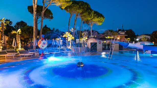 A swimming pool with water jets, lounge chairs, and a small building in the background. - Hotel Mediterranee Family & Spa Hotel (San Michele al Tagliamento)