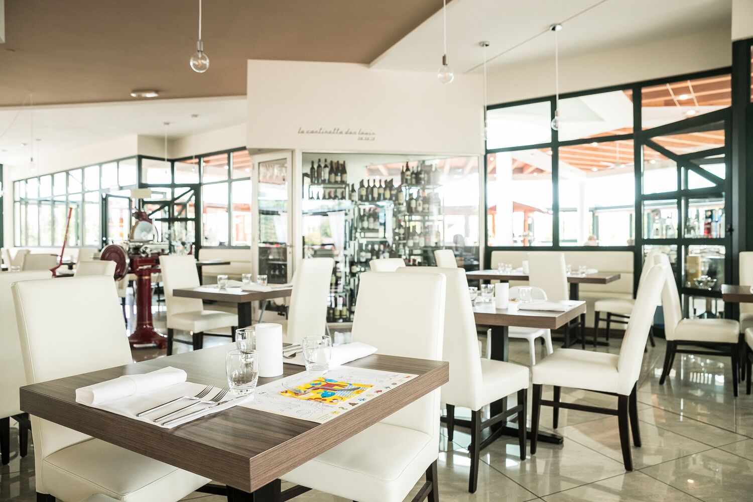 Interior moderno de restaurante com cadeiras de couro branco, mesas de madeira e um display de vinhos ao fundo.