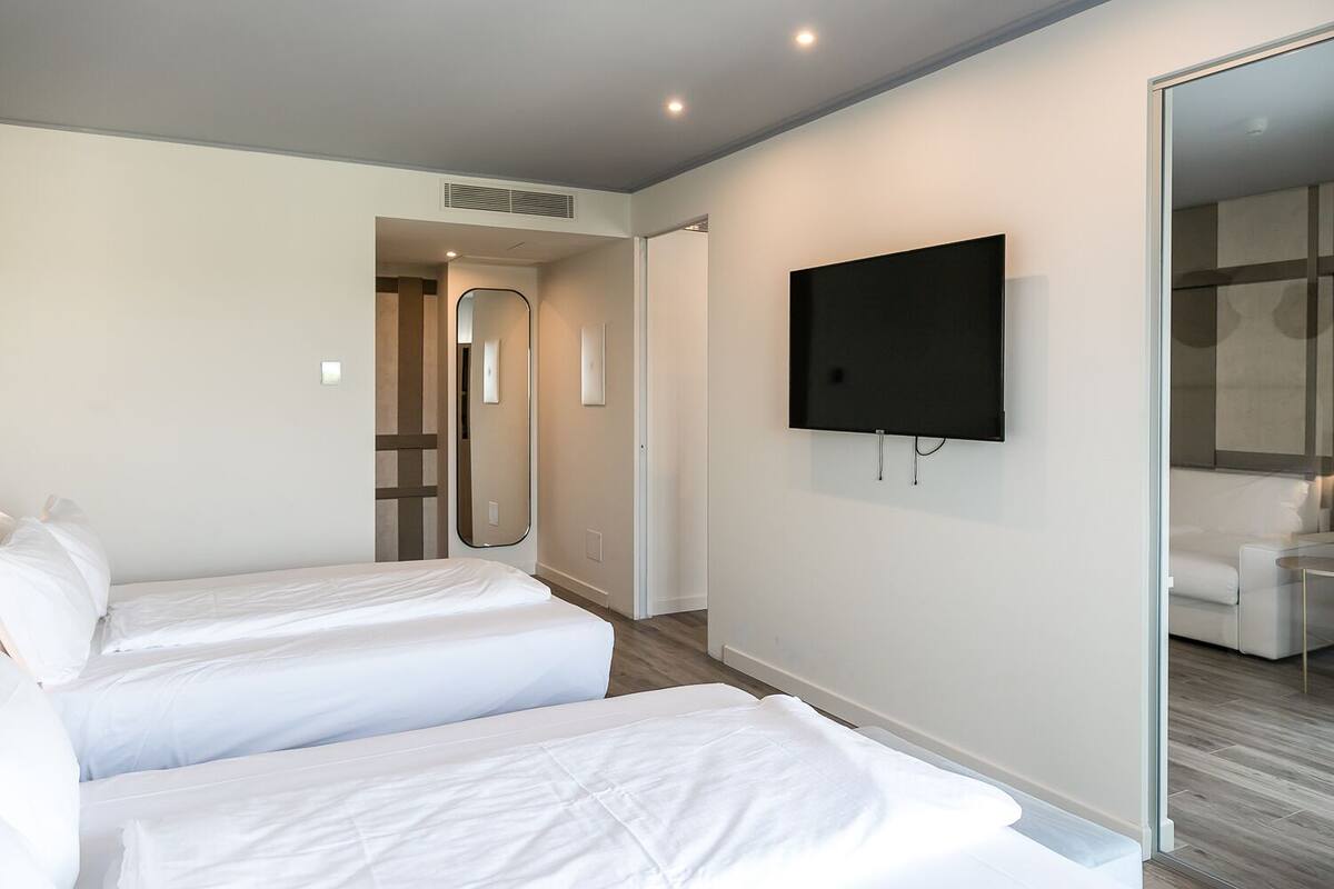 A modern hotel room with two single beds, a flat-screen TV mounted on the wall, and a glass door leading to a bathroom.
