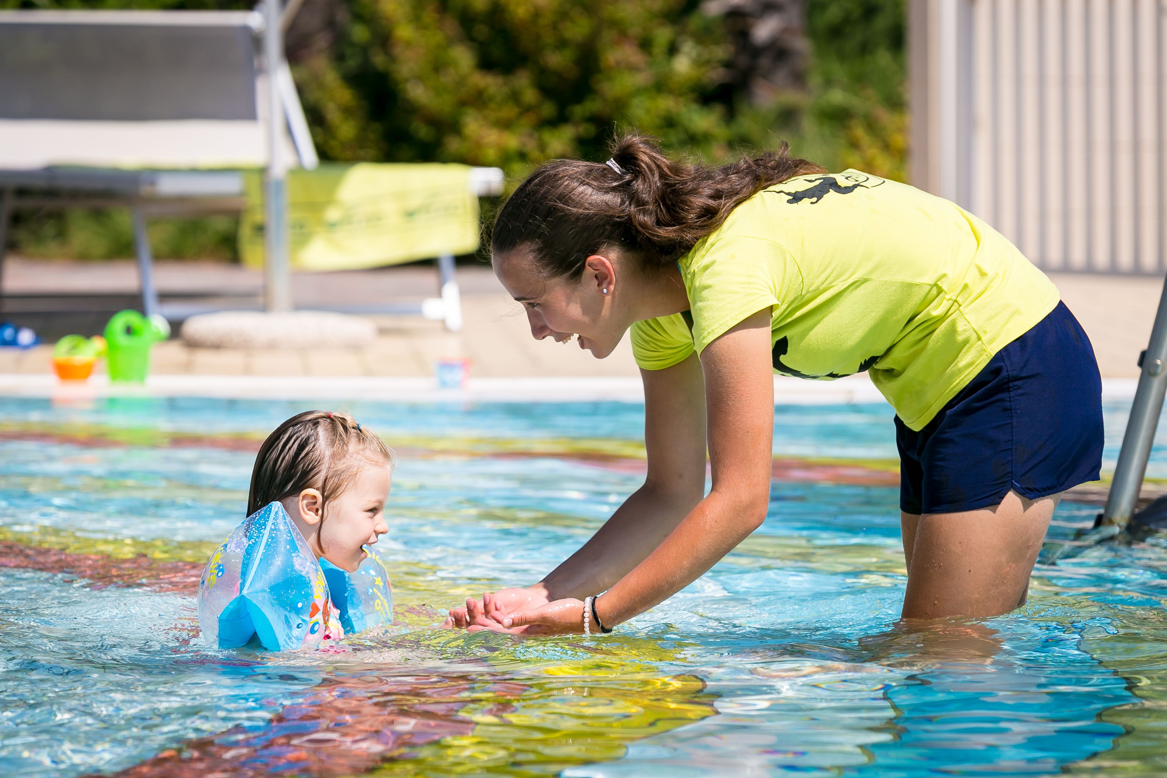 Piscina com água cristalina, uma criança usando touca de natação e um adulto auxiliando a criança.