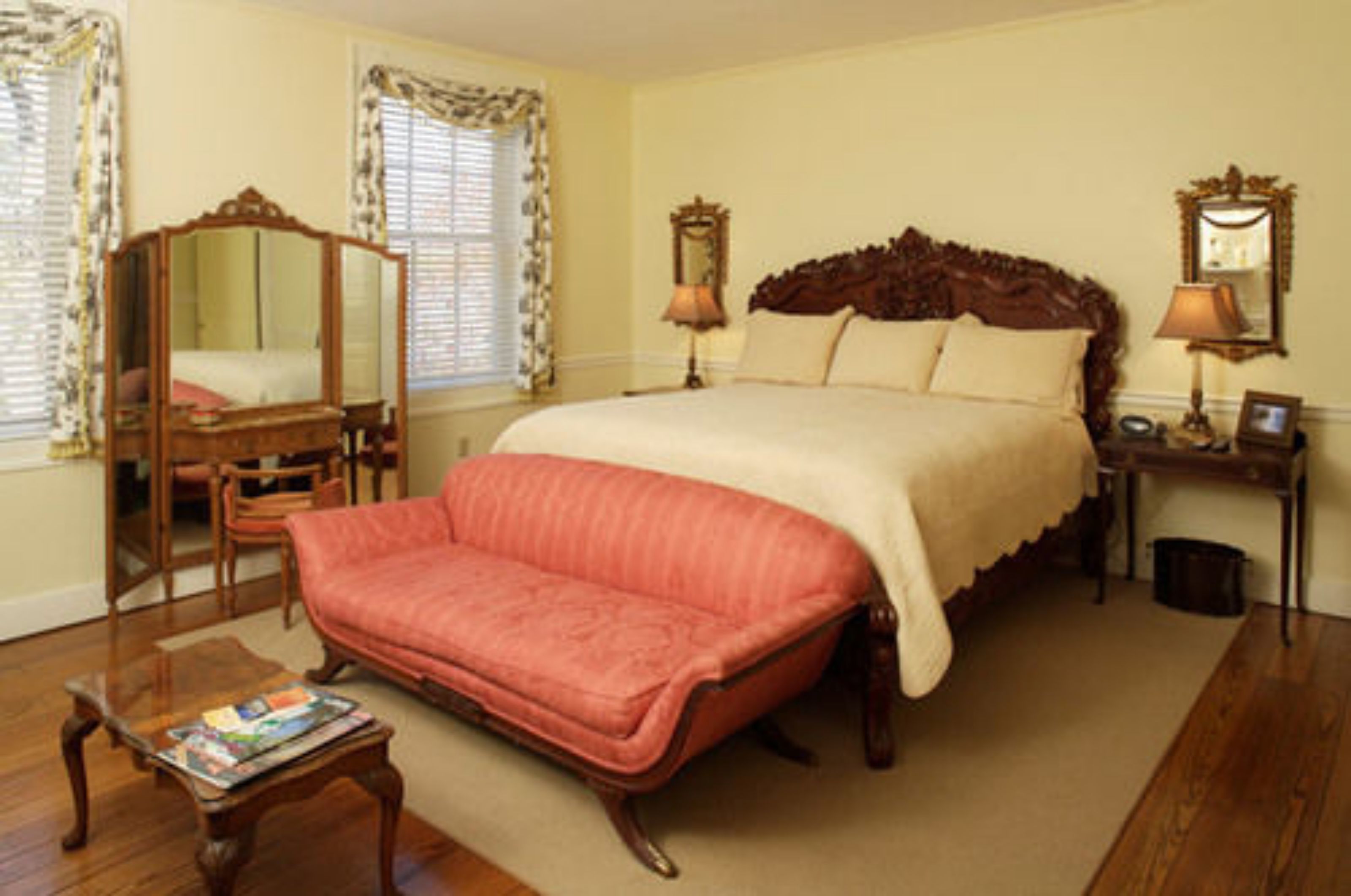 A bedroom with a large bed, a chaise lounge, a wooden nightstand with a lamp, a mirror, and a window with curtains.