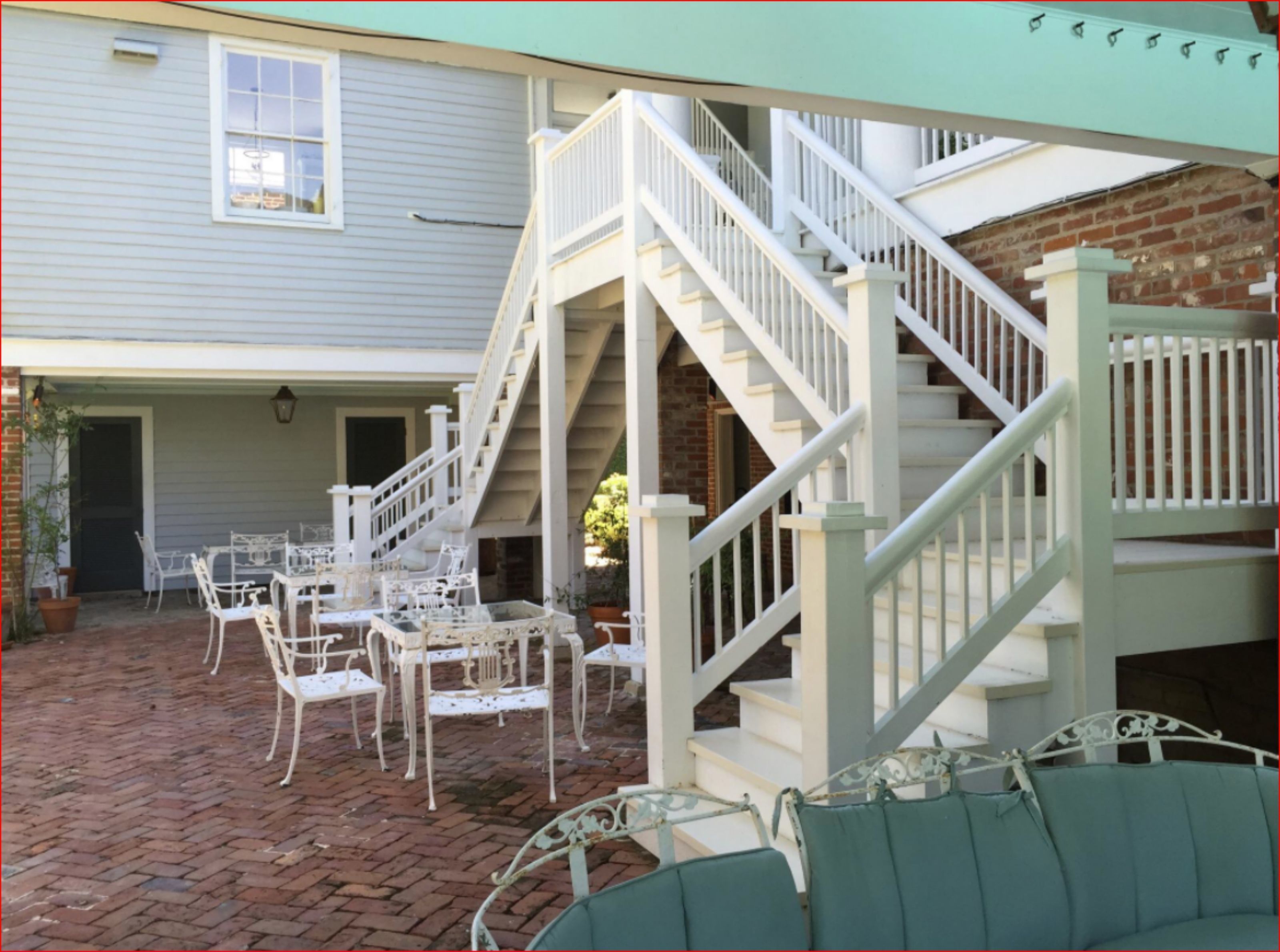 A covered patio with a brick floor, white metal furniture, and a white staircase.