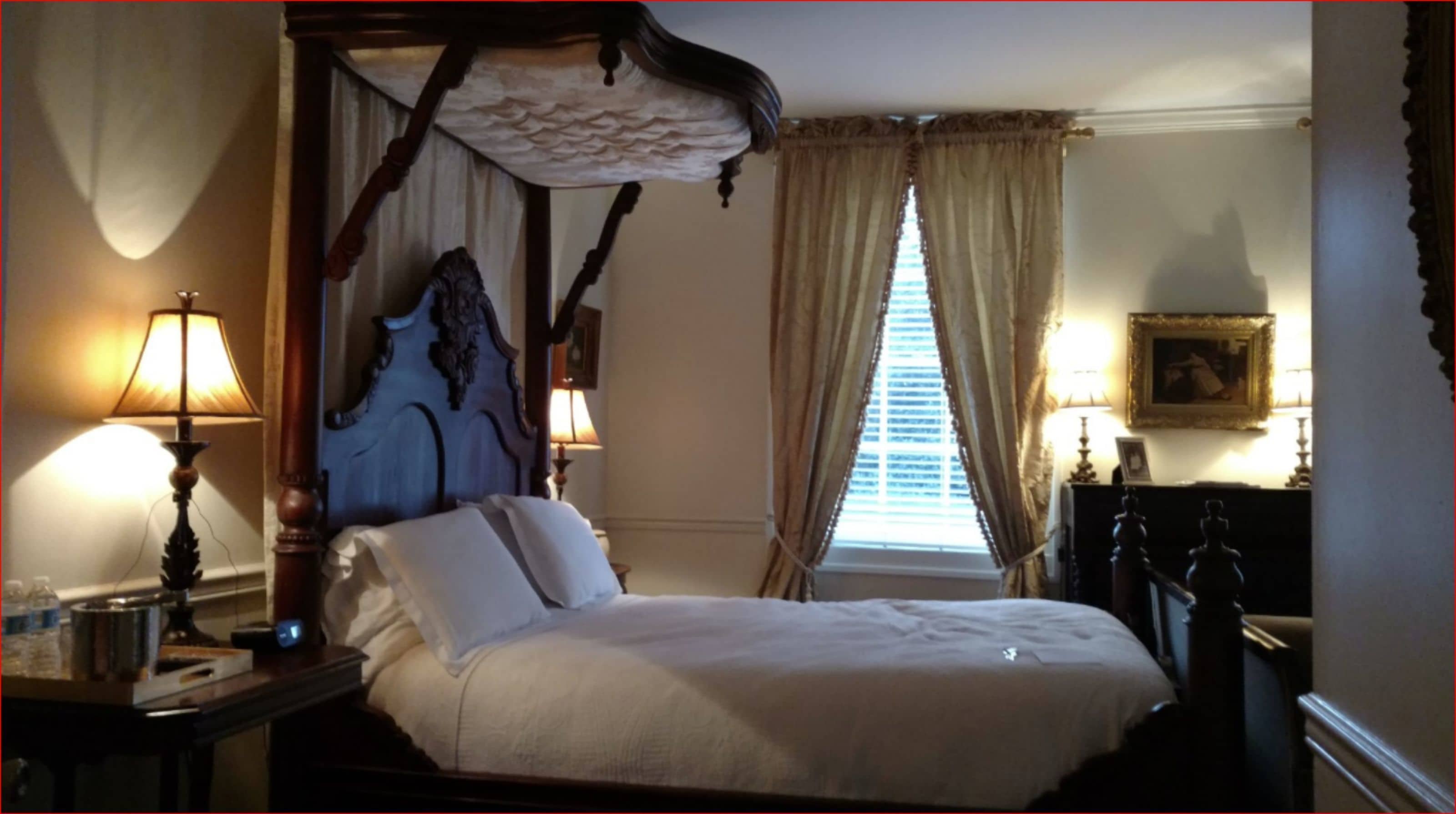 A four-poster bed with a canopy, two bedside lamps, a nightstand, a dresser, a window with curtains, and a framed picture on the wall.