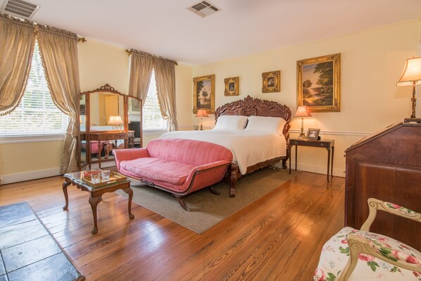 A bedroom with a large bed, a vanity with a mirror, a pink armchair, and a wooden dresser. - The Burn Bed and Breakfast (Natchez)