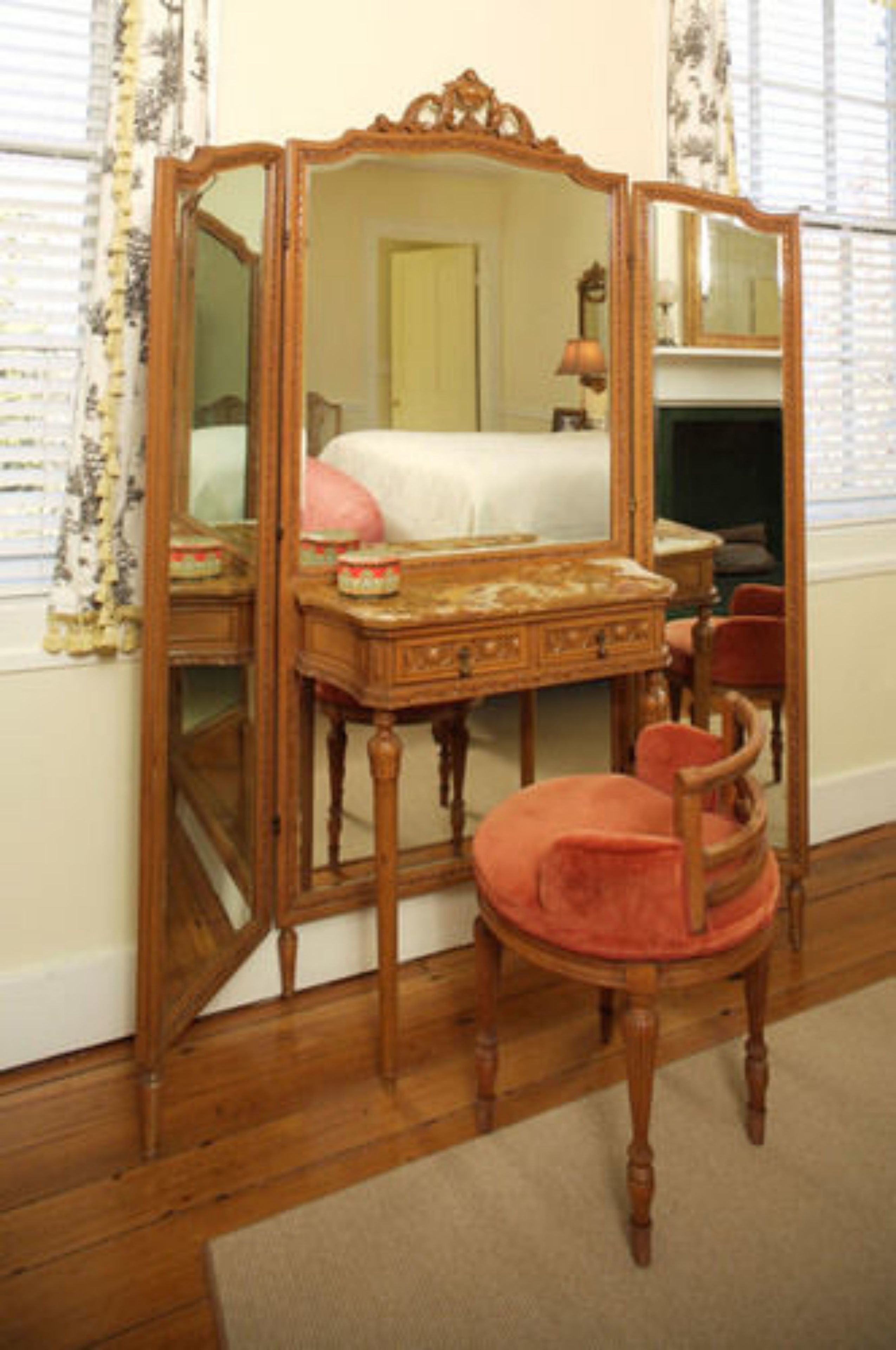 A room with a wooden vanity, a mirror, a pink chair, and a bed with white bedding.