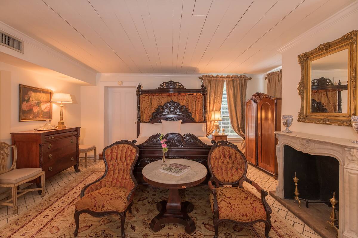 Una habitación tradicional con cama, dos sillones, una mesa redonda, un aparador, una lámpara, un cuadro y una chimenea.