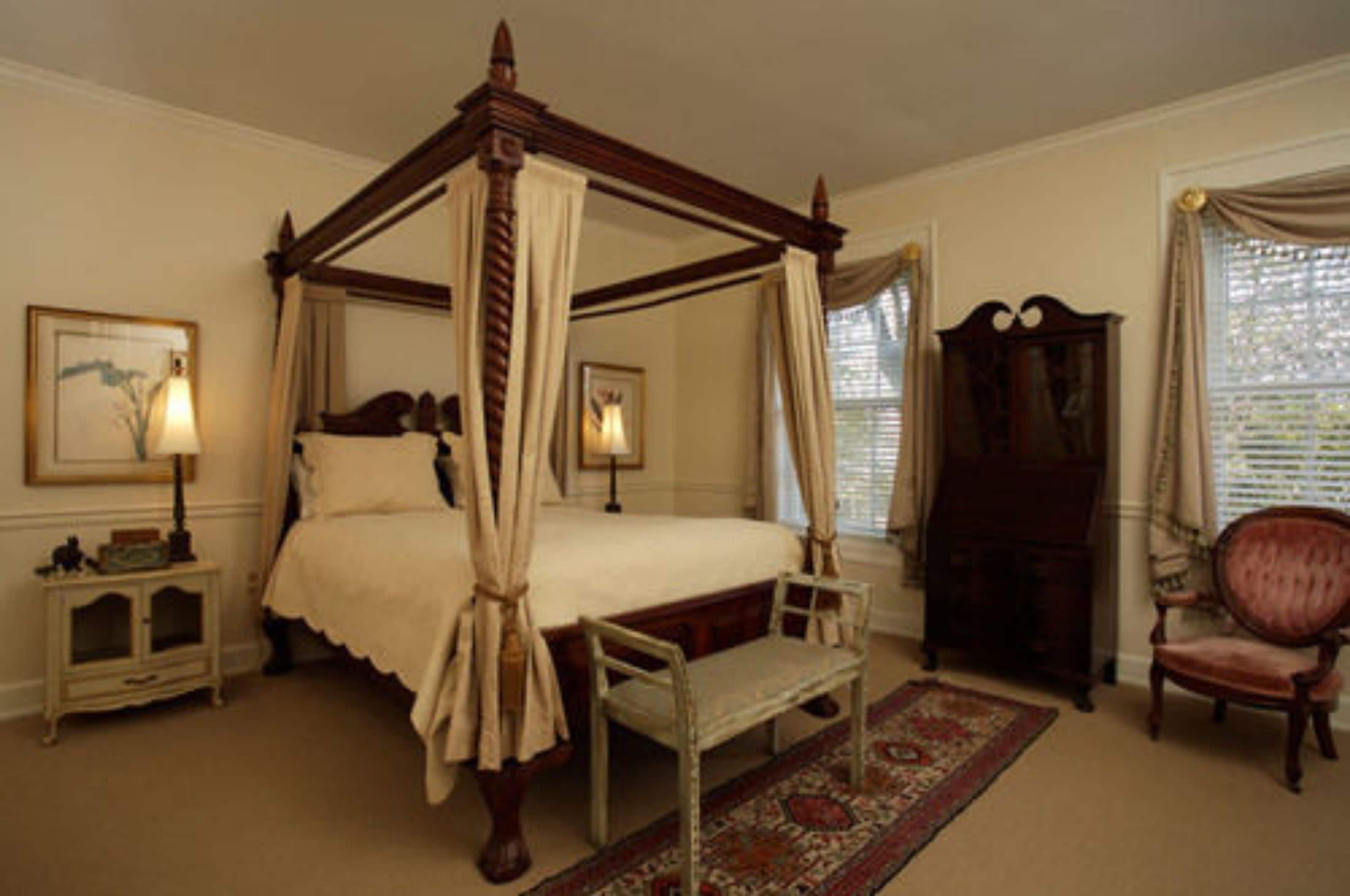 A four-poster canopy bed with a bench and a chair, a wooden cabinet, and a framed picture on the wall.