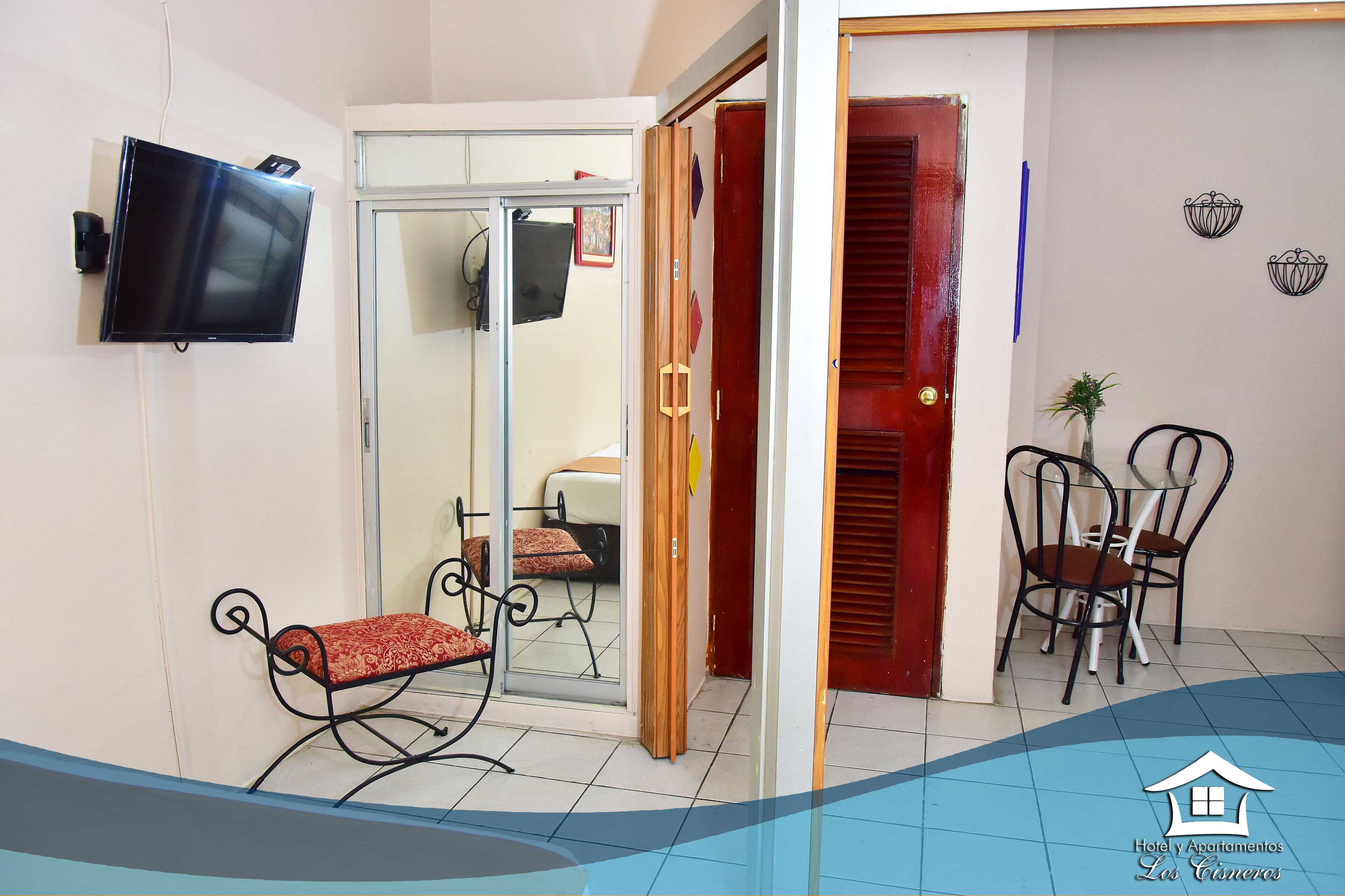 A hotel room with a TV mounted on the wall, a sliding glass door leading to a balcony, and a small seating area with a bench and chairs.