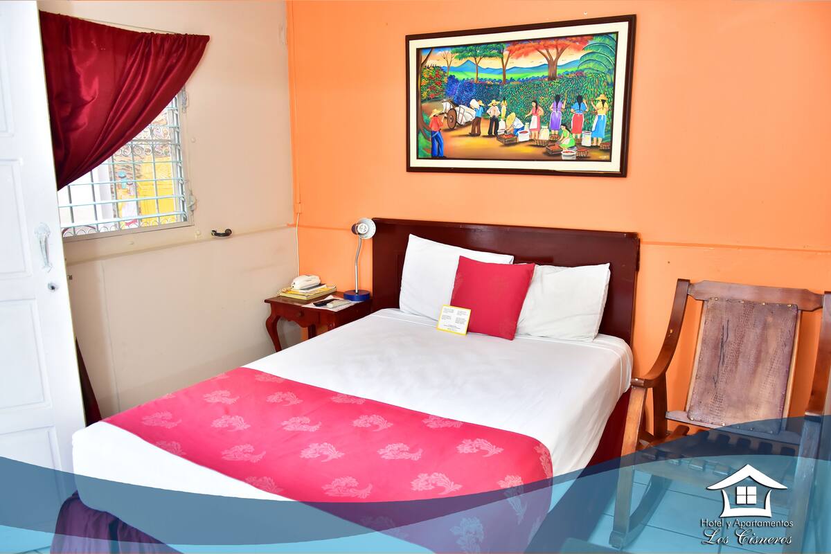 A hotel room with a bed, a nightstand, a chair, a window with curtains, and a painting on the wall.