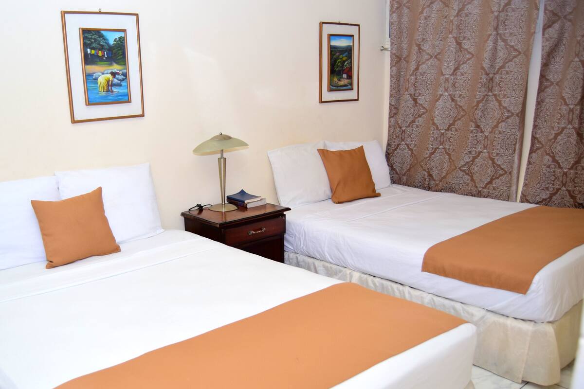Two single beds with white linens and orange throw pillows, a wooden nightstand with a lamp, and two framed pictures on the wall.