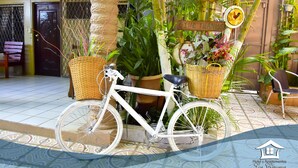 Una bicicleta blanca estacionada en un espacio exterior cuidado, con plantas en macetas y un cartel de bienvenida.