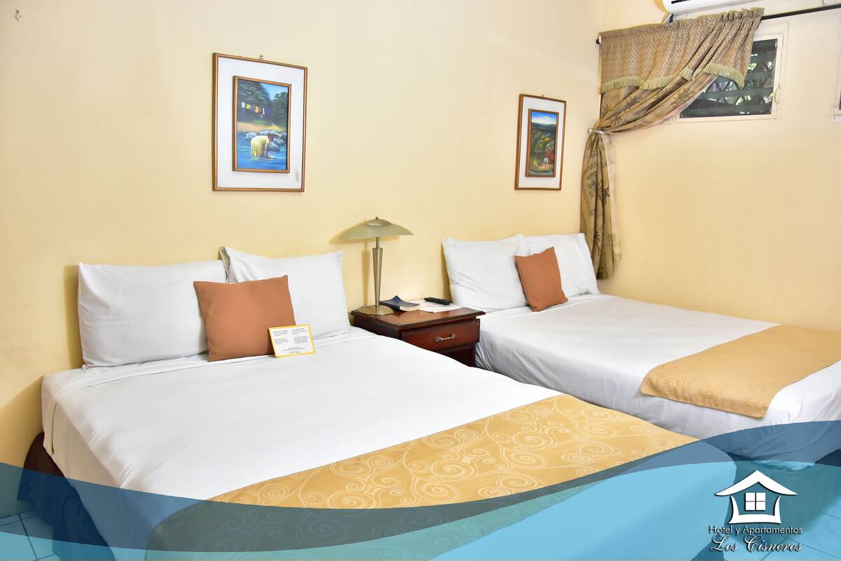 A hotel room with two beds, a nightstand, a lamp, and framed pictures on the wall.