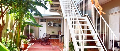 A narrow outdoor corridor with a red carpet, a white staircase, and potted plants.