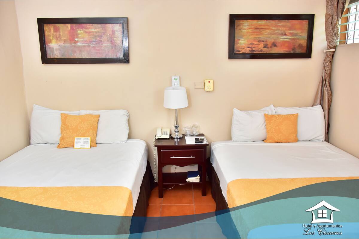 A hotel room with two beds, a nightstand with a lamp, a phone, and a small table.