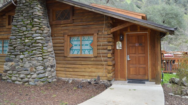 A rustic log cabin with a stone chimney, wooden door, and a window with a geometric pattern. - Red Bud Lodge (El Portal)