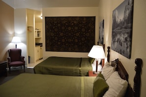 A hotel room with two beds, a chair, a lamp, a wardrobe, and a tapestry.