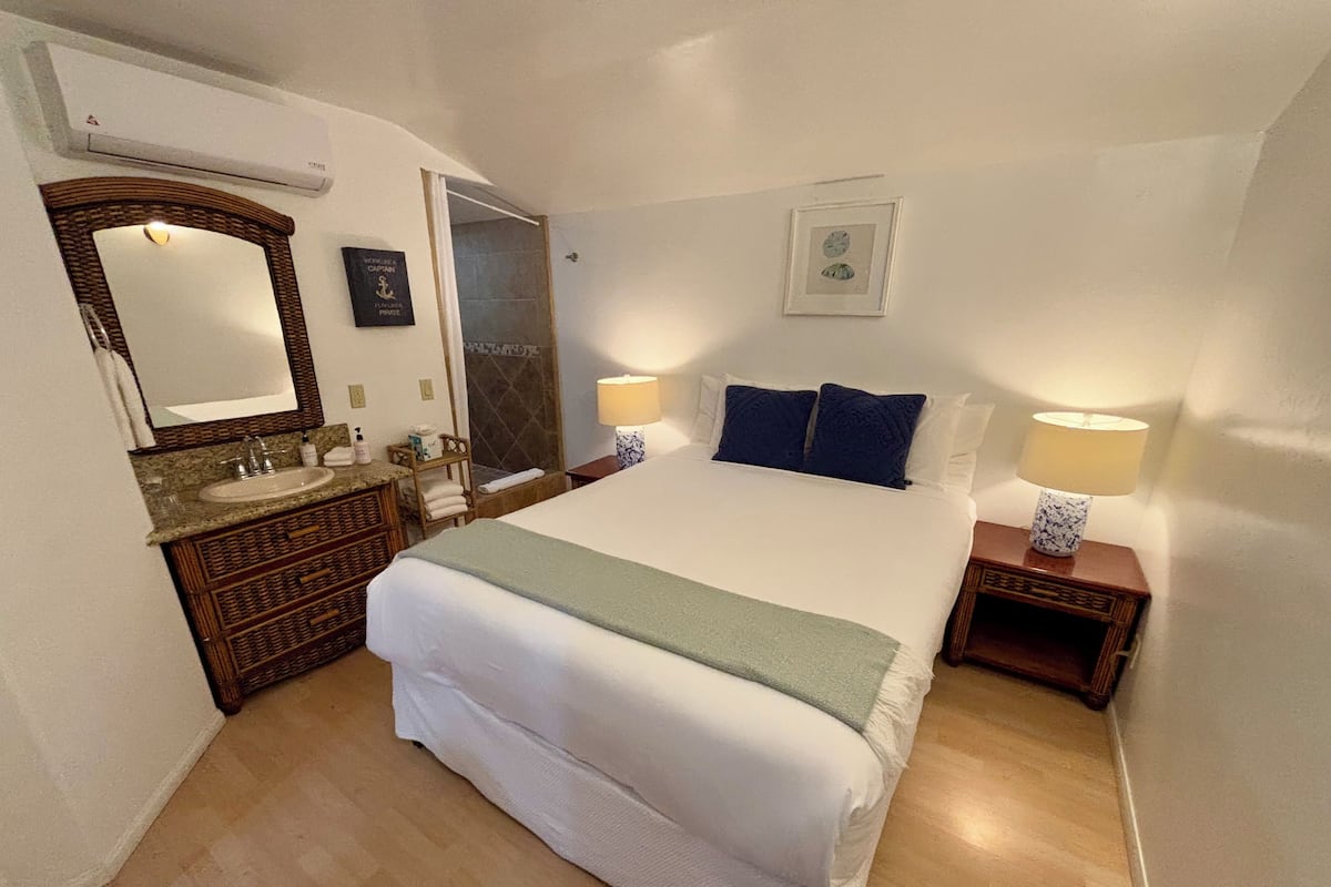 Quarto de hotel com uma cama grande, banheiro com pia e espelho, e duas mesas de cabeceira com abajures.