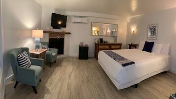 A hotel room with a large bed, a television mounted on the wall, a fireplace, a desk with a lamp, and a dresser.