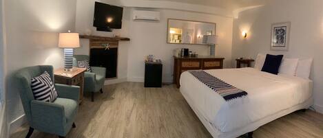 A hotel room with a large bed, a television mounted on the wall, a fireplace, a desk with a lamp, and a dresser.