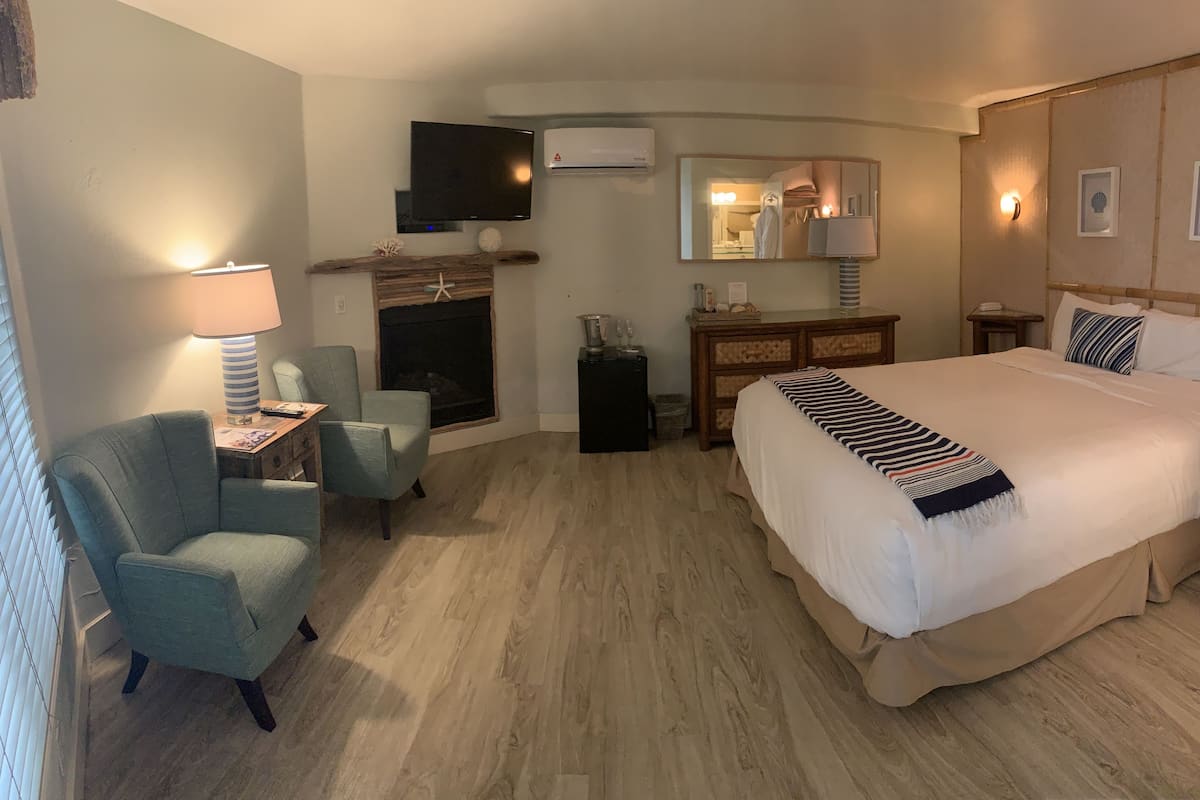 Quarto de hotel com uma cama grande, escrivaninha com luminária, cadeira, lareira, televisão e espelho.
