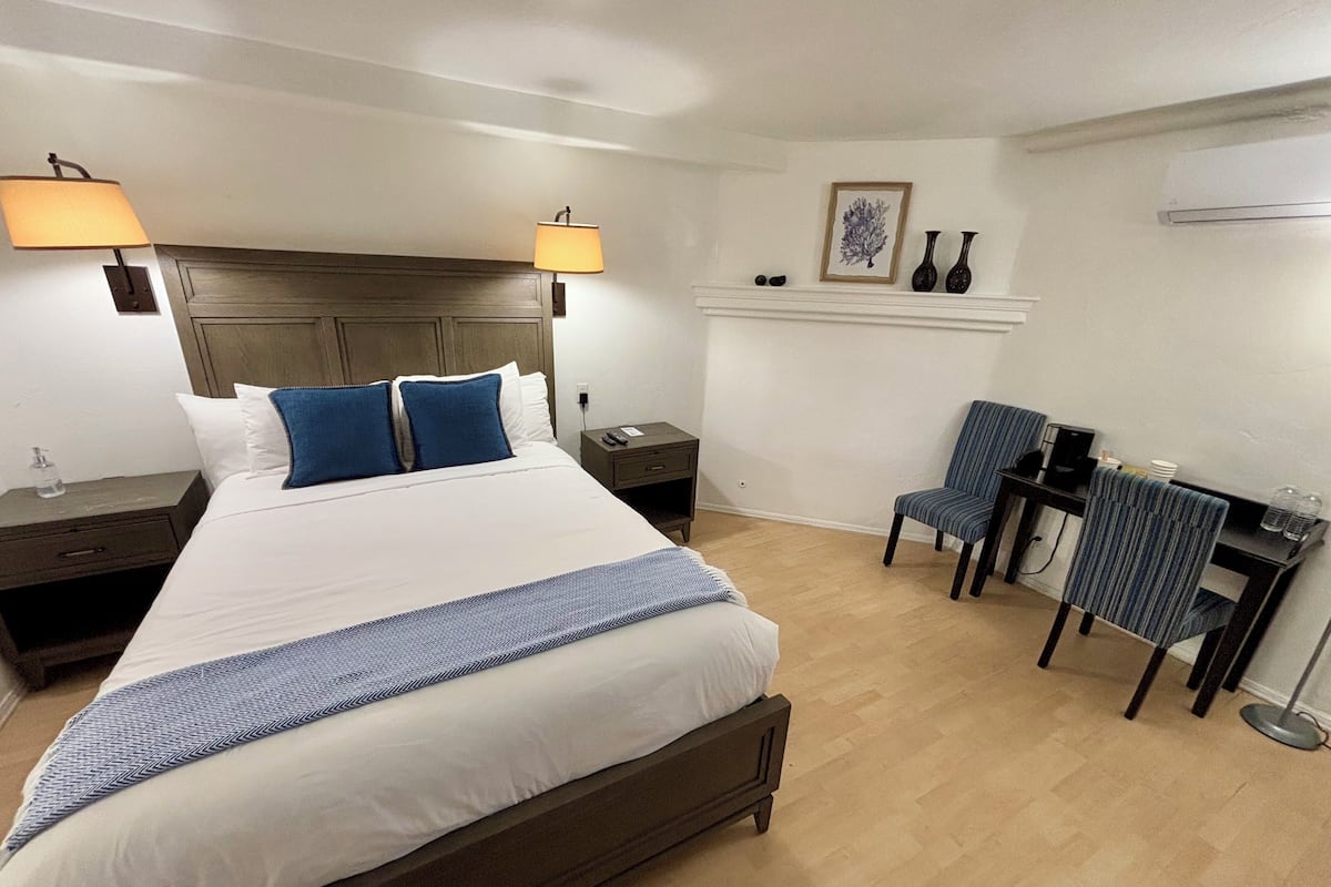 Quarto de hotel com uma cama grande, duas mesas de cabeceira, uma escrivaninha com cadeira e uma pequena estante com objetos decorativos.