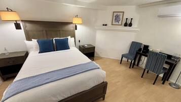 A hotel room with a large bed, two bedside tables, a desk with a chair, and a small shelf with decorative items.