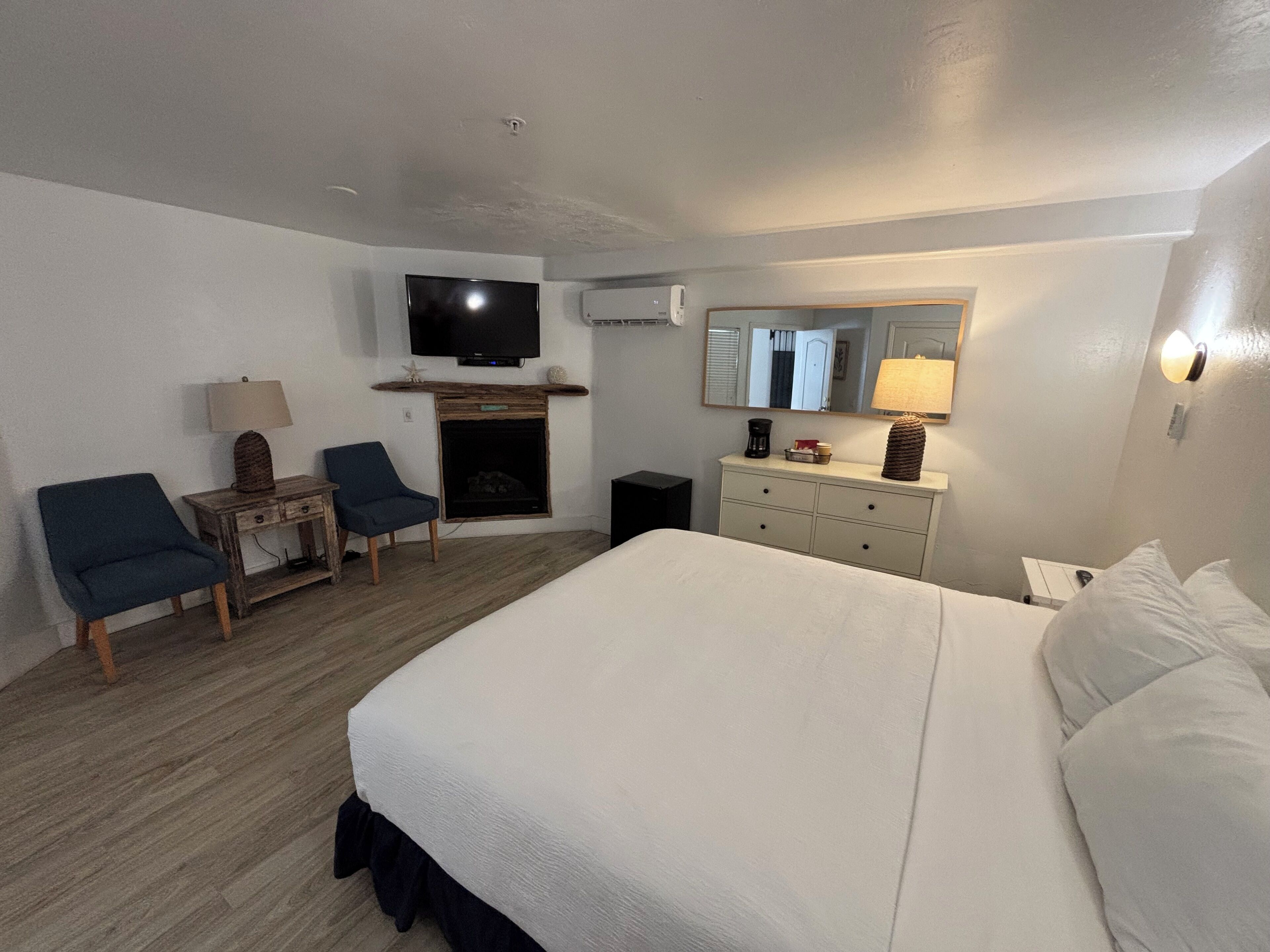 A hotel room with a large bed, two armchairs, a small table with a lamp, a fireplace, a television, and a mirror.