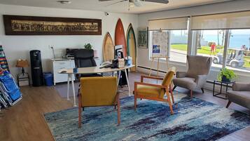 A room with a desk, two chairs, a surfboard, a beach-themed poster, and a view of the beach through large windows.