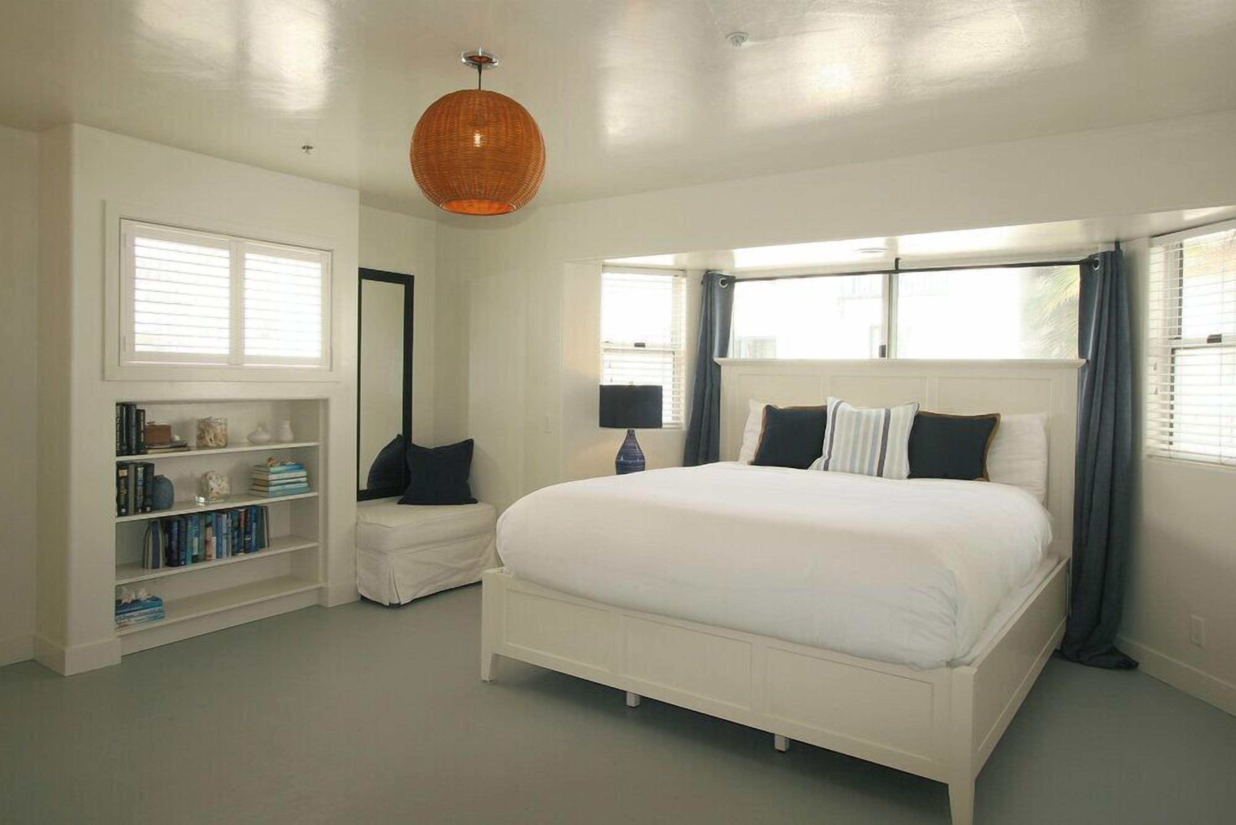 A bedroom with a white bed, a bookshelf, a mirror, and a hanging light fixture.