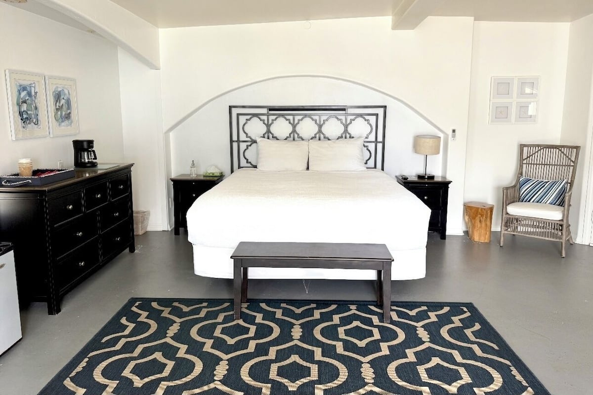 Quarto com uma cama grande, cómoda de madeira escura, mesa de cabeceira, cadeira e tapete estampado.
