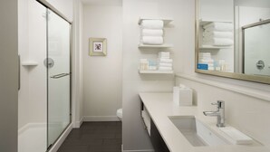 Un baño moderno con cabina de ducha de vidrio, tocador blanco con lavamanos y espejo, y estantes con toallas dobladas con cuidado.
